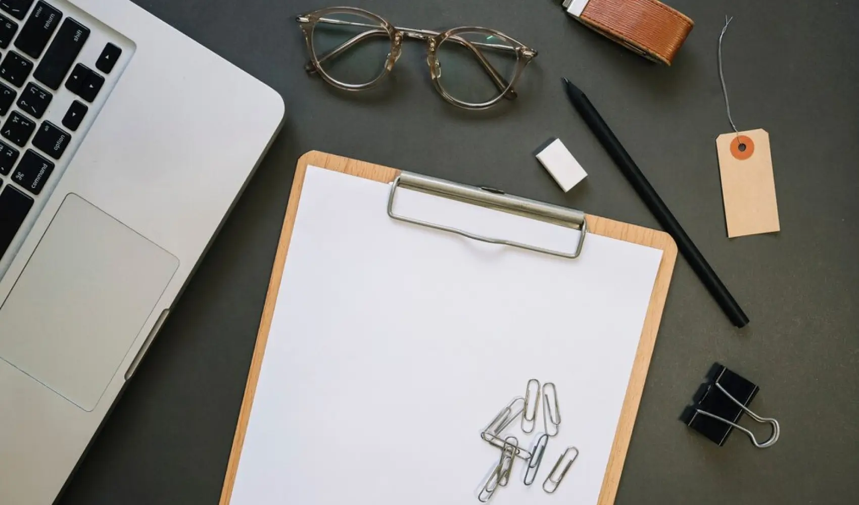 Clipboard with blank paper, paperclips, pen, glasses, eraser, and notebook on a desk, representing tools for employee onboarding checklist and organization.